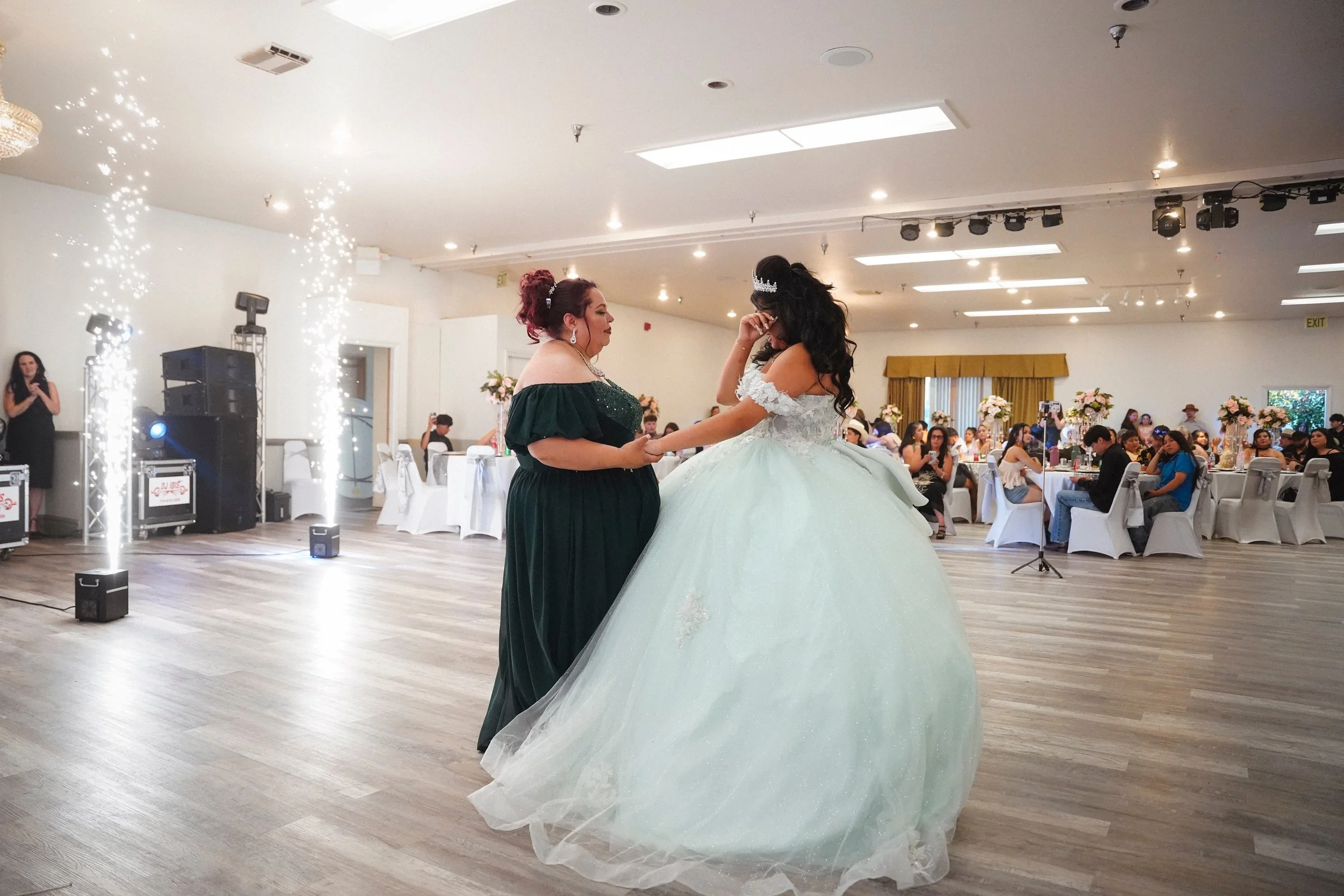 A bride in a white wedding gown and tiara dancing with a woman in an off-the-shoulder dark green gown at a wedding reception. Guests are seated at decorated tables in the background.