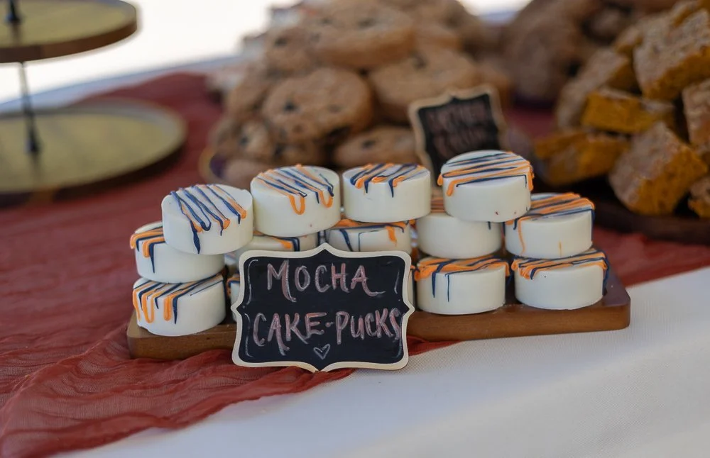 Display of mocha cake pucks with white icing and orange and blue drizzle, labeled with a chalkboard sign.
