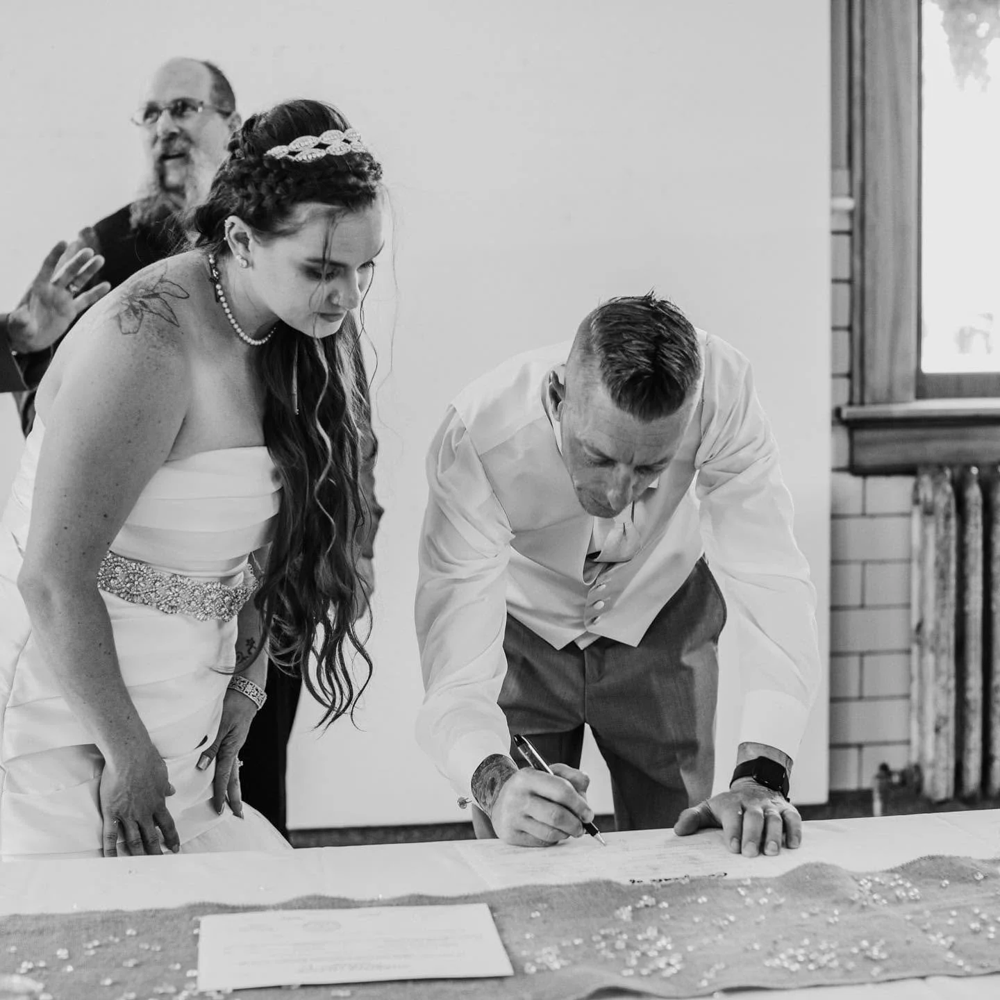 A wedding ceremony where a bride in a strapless white gown and a necklace signs a document while a man in a white shirt signs next to her. A woman with long curly hair, dressed in wedding attire, observes, with a man in glasses and a beard in the bac