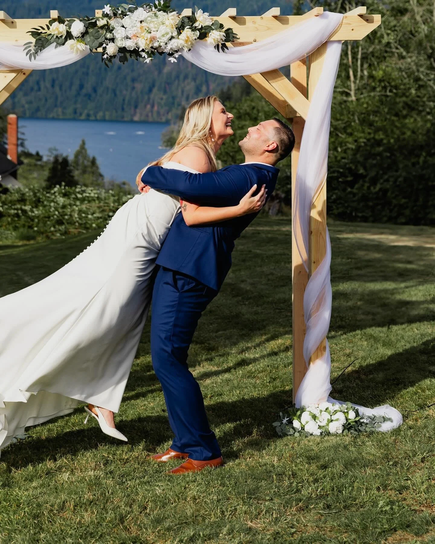 A newlywed couple appears to be dancing at an outdoor wedding ceremony under a wooden arch decorated with white flowers and sheer fabric, with a landscape of water and forested hills in the background.