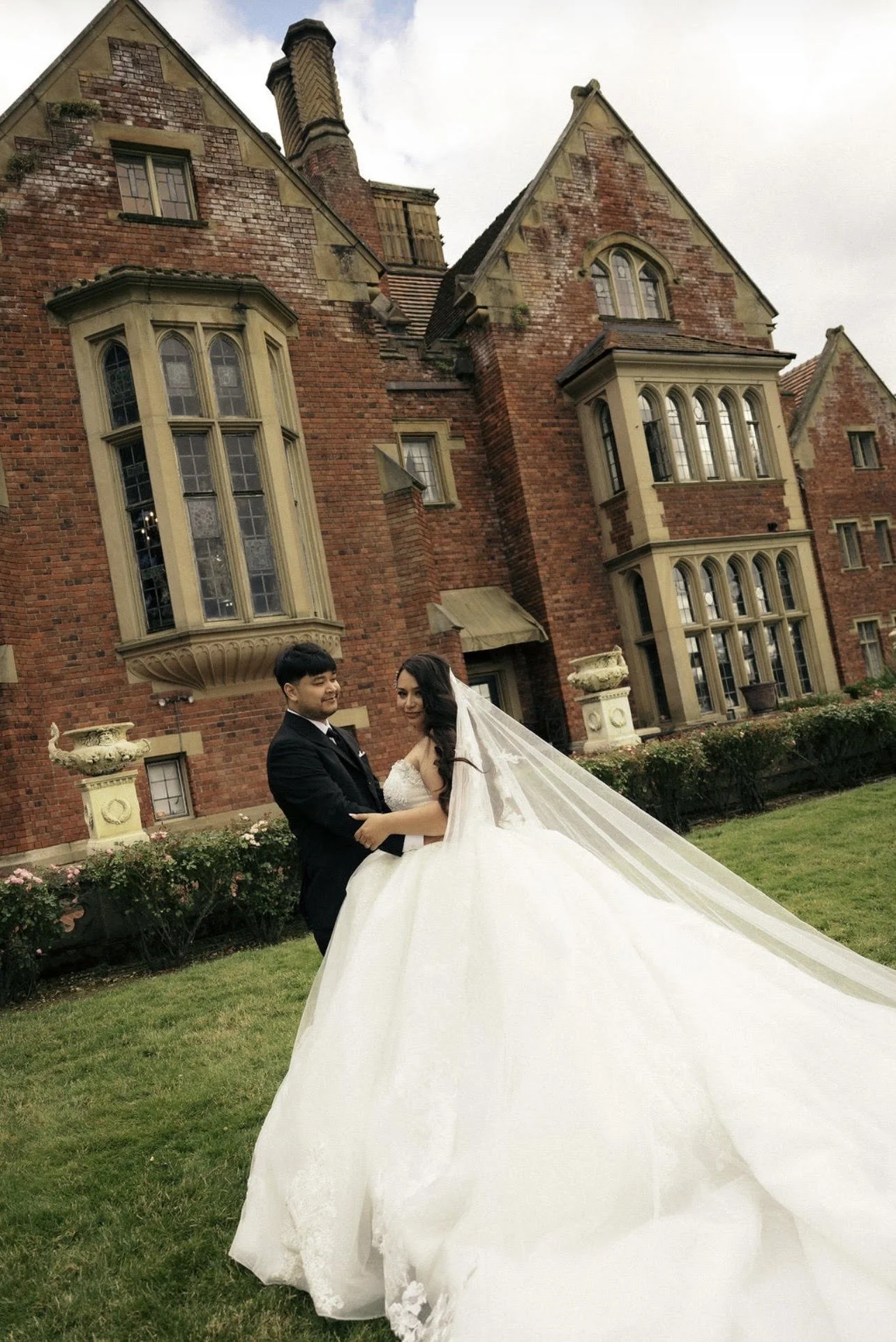 A bride and groom standing outdoors on a grassy area in front of a large brick mansion with multiple windows, columns, and decorative stone features. The bride is wearing a white wedding gown with a long train and veil, while the groom is dressed in 
