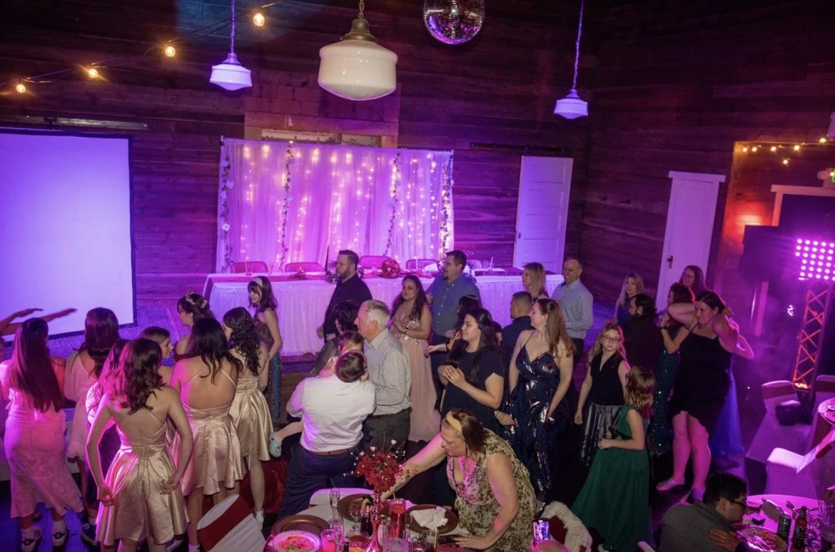 Indoor celebration with people dancing and socializing, wooden walls and ceiling, decorated with string lights and a purple backdrop, with a table of decorations and flowers in the foreground.