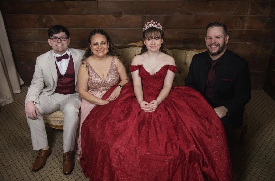 A young woman wearing a red ball gown and tiara sitting on a vintage sofa, flanked by a man in a black suit, a woman in a pink dress, and a young man in a cream suit with a burgundy vest and bowtie, all smiling for a group photo.
