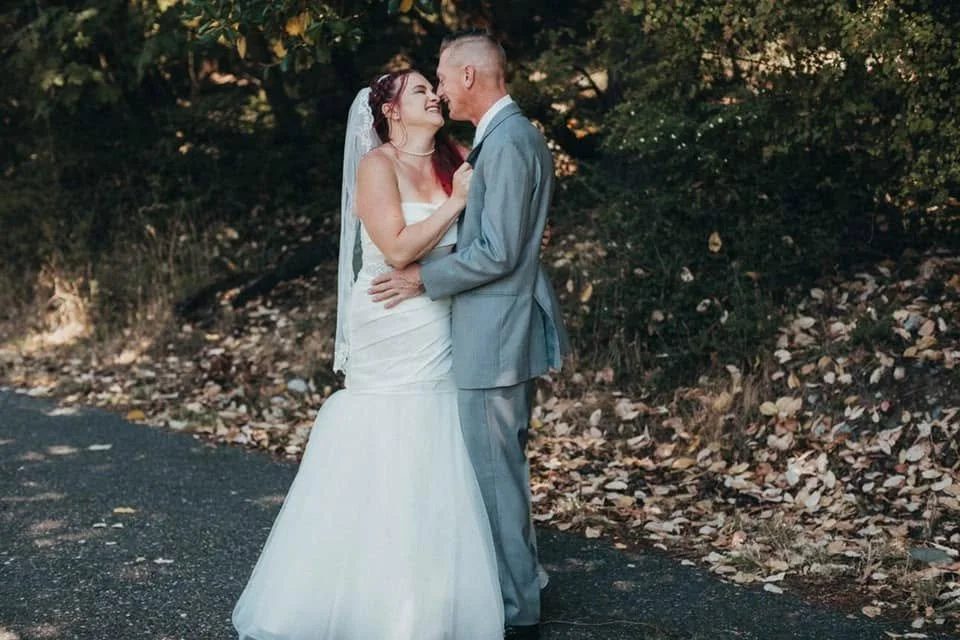 A bride and groom in wedding attire are smiling and close together outdoors, surrounded by trees and fallen leaves.