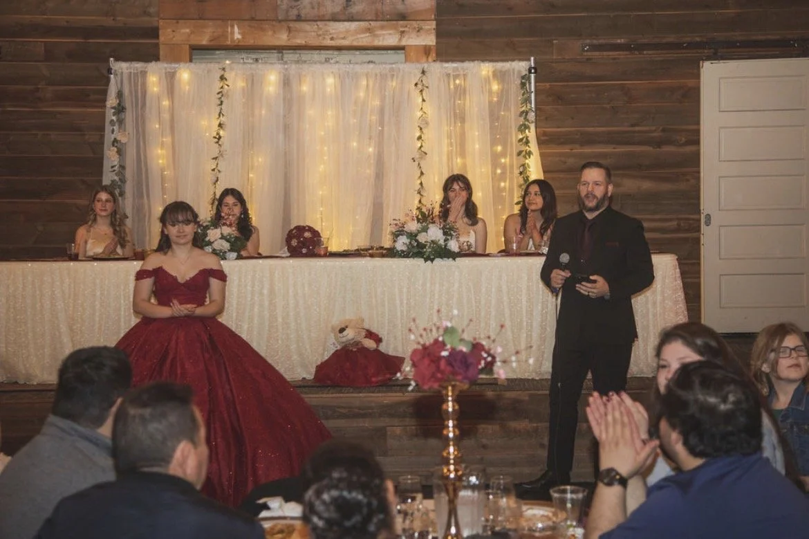 A wedding reception with a bride in a red gown standing in front of a seated head table, decorated with flowers and fairy lights, while a man in a black suit holds a microphone and gives a speech, surrounded by guests clapping and watching.