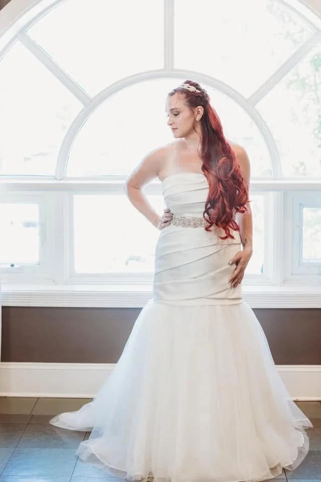 A woman in a strapless white wedding gown with a bejeweled waistline, standing in front of a large arched window with light streaming in.
