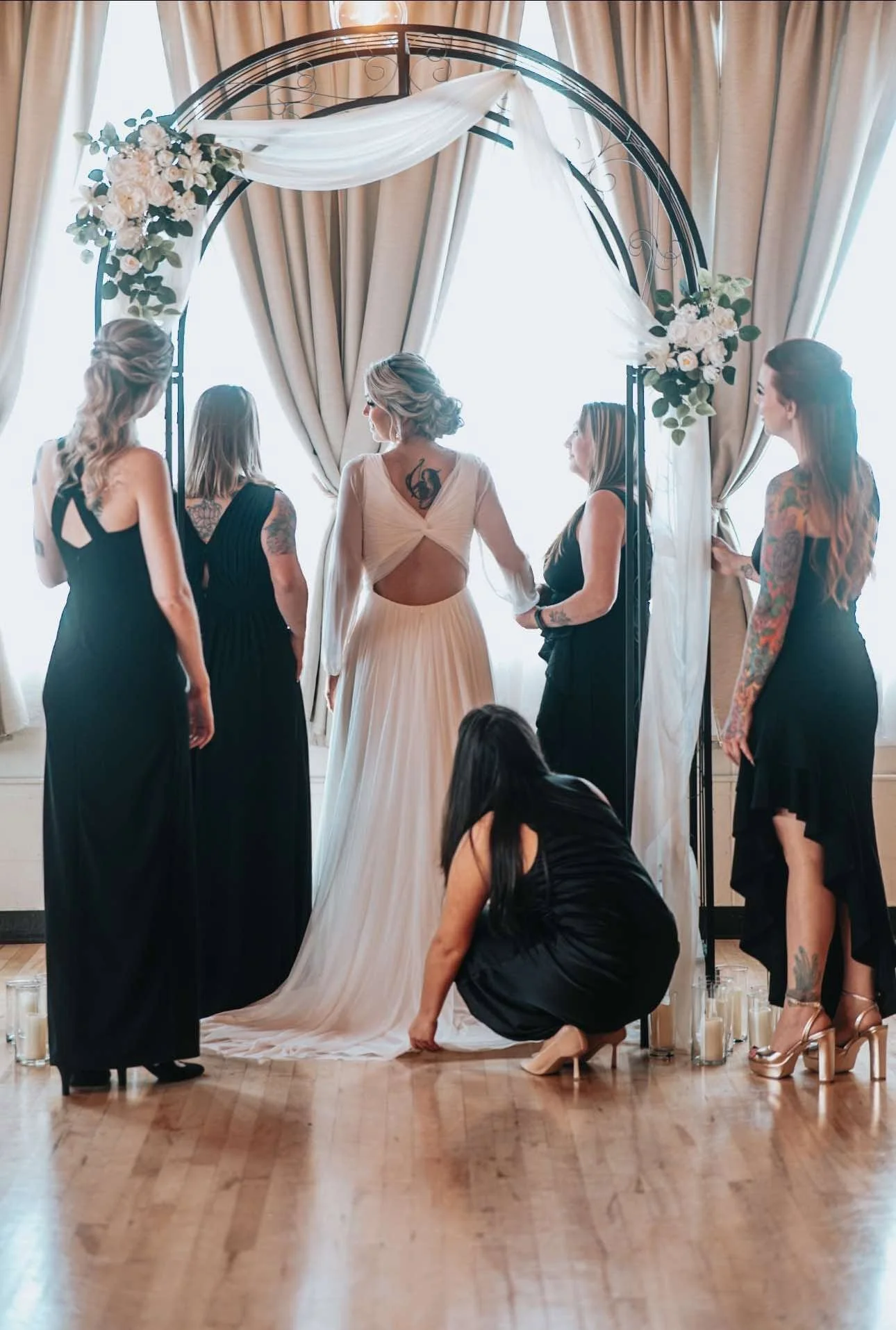 A bride in a white wedding dress standing under a floral wedding arch with her bridesmaids during a wedding ceremony, with candles on the floor.