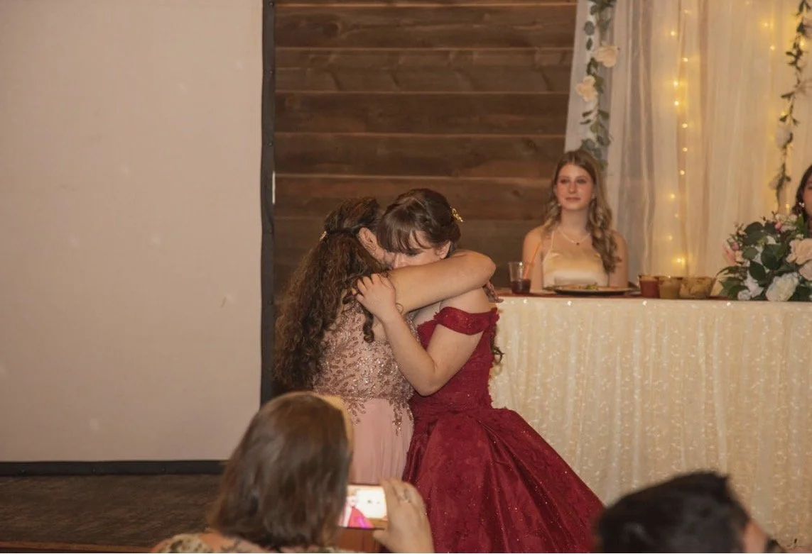Two young girls hugging each other at a celebration or formal event, with one in a pink dress and the other in a red dress. In the background, there is a woman seated at a table decorated with lights and flowers.