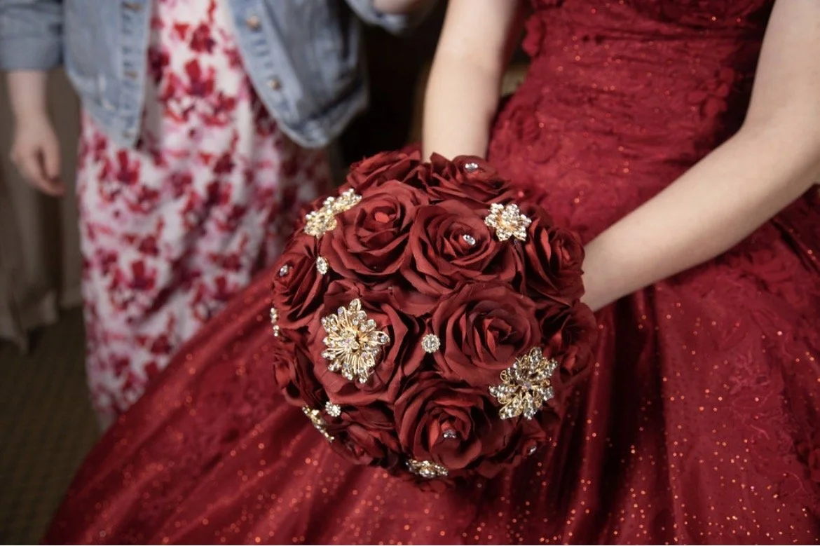A woman in a red dress holding a bouquet of red roses decorated with gold and pearl jewelry.