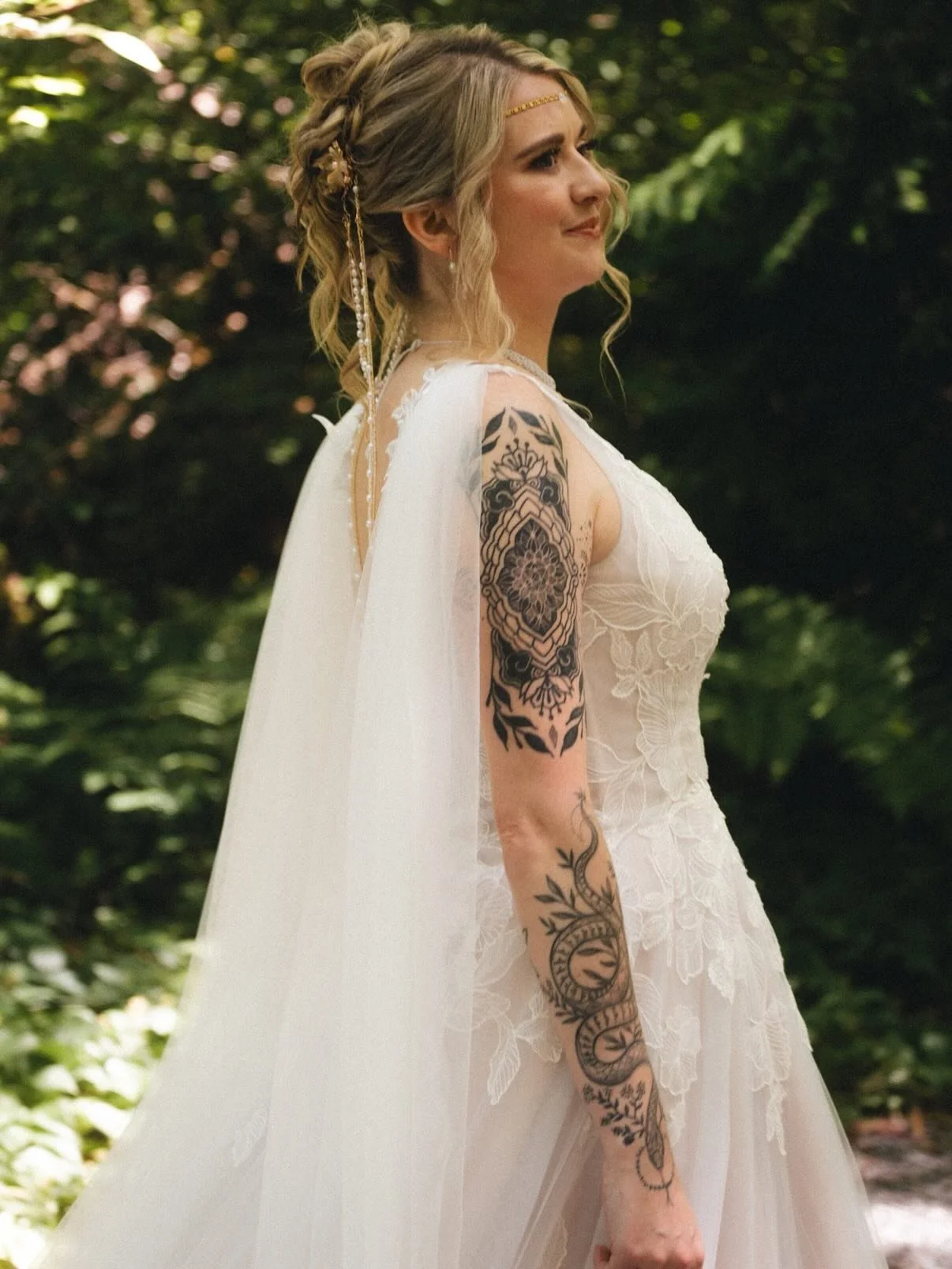 A woman in a white lace dress with a long veil stands outdoors with a lush green background. She has styled blonde hair with braids and hair accessories, and visible tattoos on her arm.