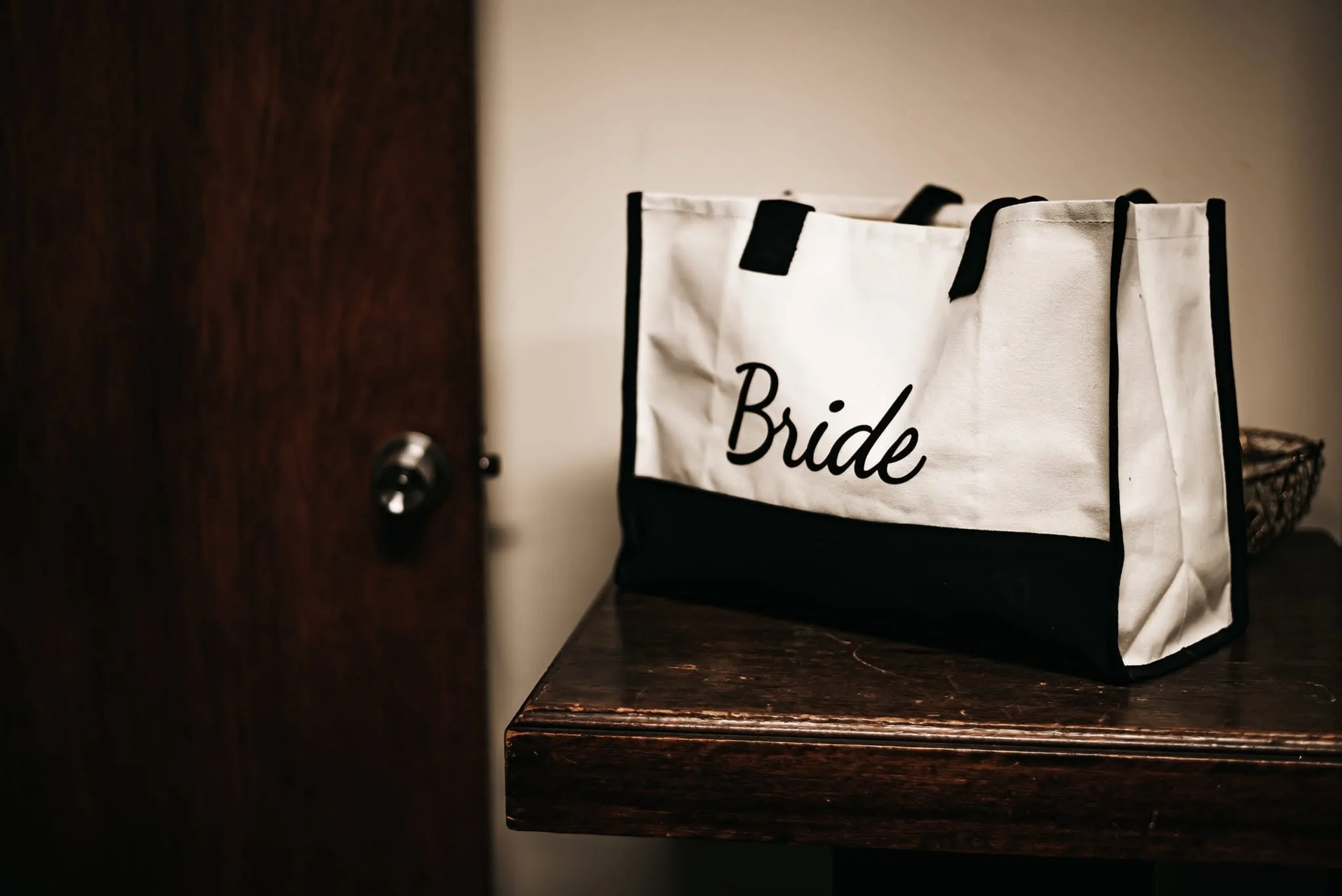A white fabric bag with black trim and handles labeled 'Bride' sitting on a wooden surface next to a dark wooden door.