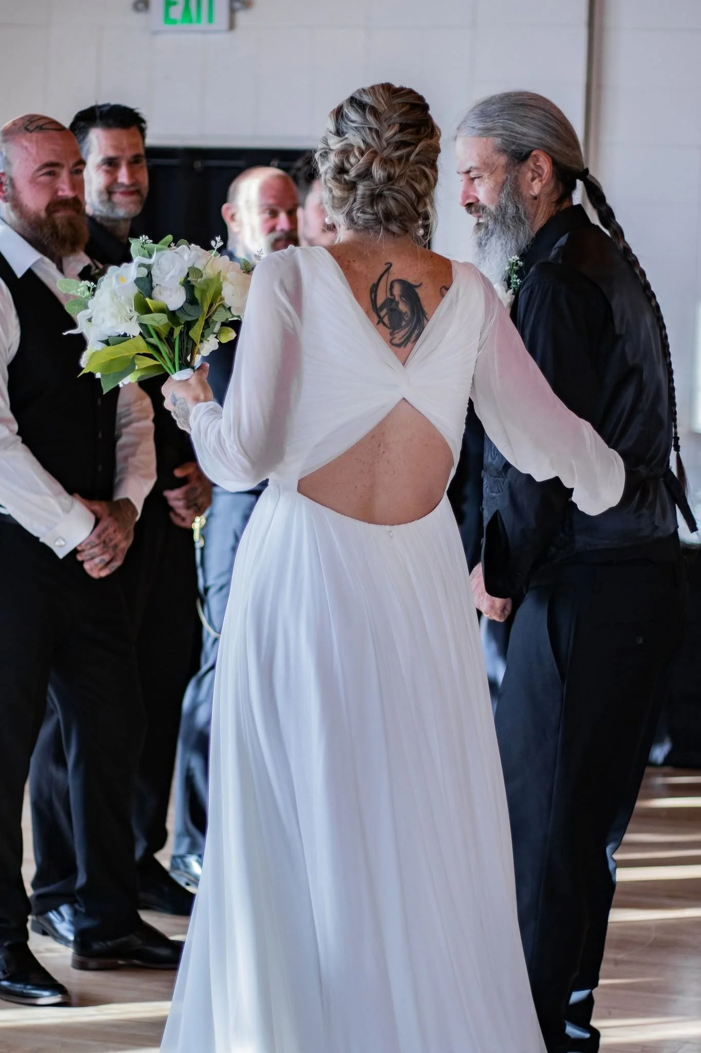 Two women with gray hair and long gray hair with a braid, wearing white and black dresses, are holding hands and dancing at a celebration. One woman is holding a bouquet of white flowers. Several men are watching and smiling in the background, dresse