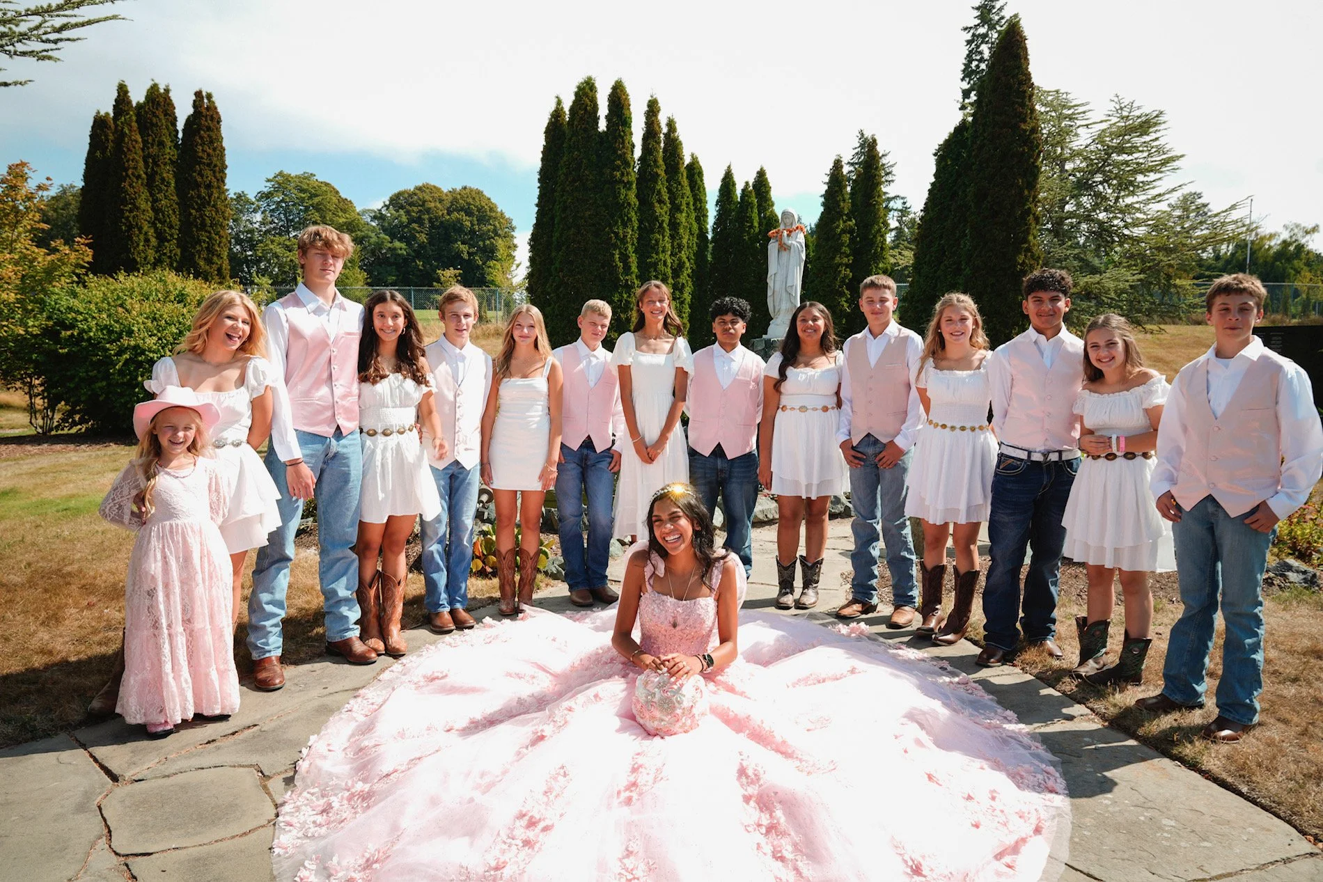 A group of children and teenagers dressed in white and pink, posing outdoors for a photo. There is a young girl in the front wearing a pink dress with a matching flower crown, seated on the ground with a large pink and white gown spread around her. T
