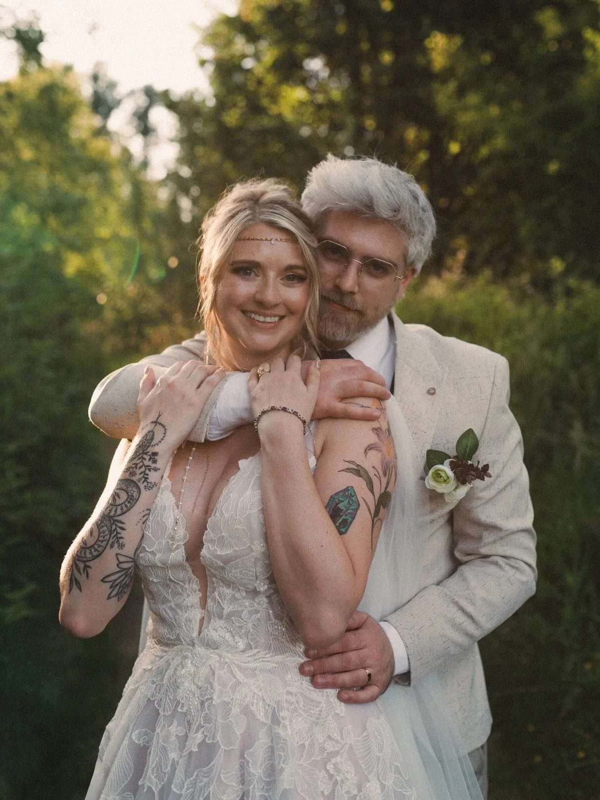 A bride and groom standing together outdoors, embracing each other, with trees and sunlight in the background.