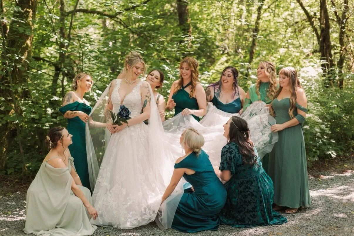 A bride in a white wedding gown holding a blue flower surrounded by bridesmaids in teal dresses outdoors in a wooded area.