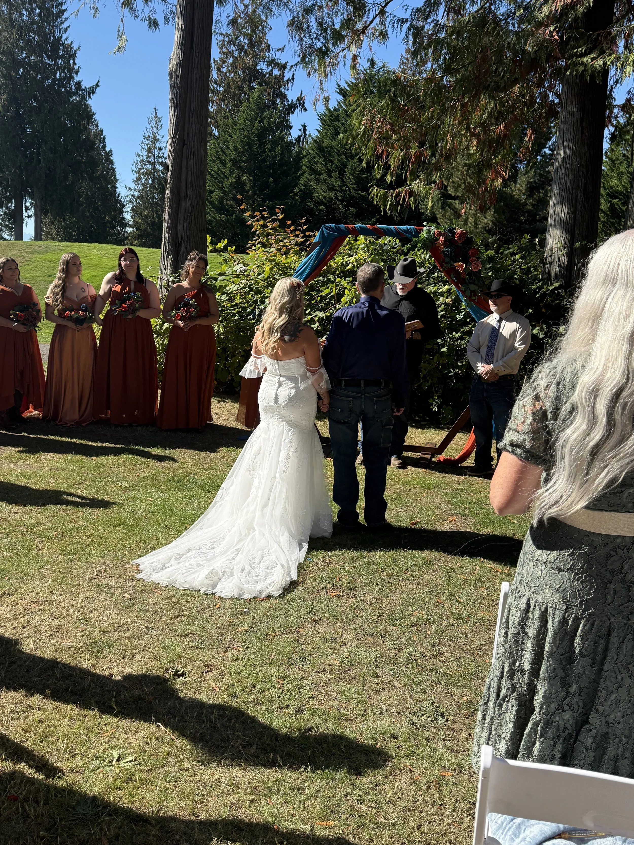 A wedding ceremony outdoors with a bride and groom standing in front of a decorated arch, surrounded by bridesmaids and guests in a scenic forest setting on a sunny day.