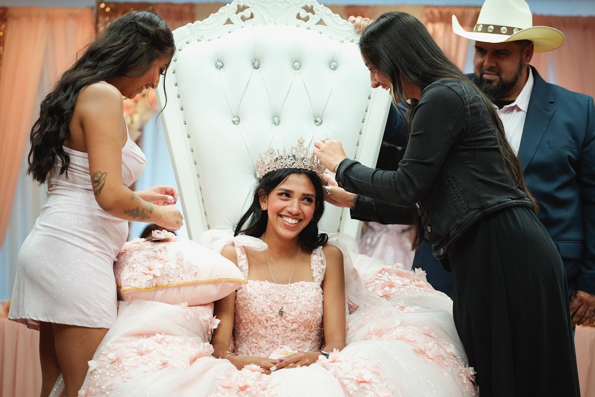 A young woman in a fancy pink dress sitting on a bed with floral decorations, smiling and wearing a crown, being crowned by two women and a man in a hat during a celebration, possibly a quinceañera or similar event.