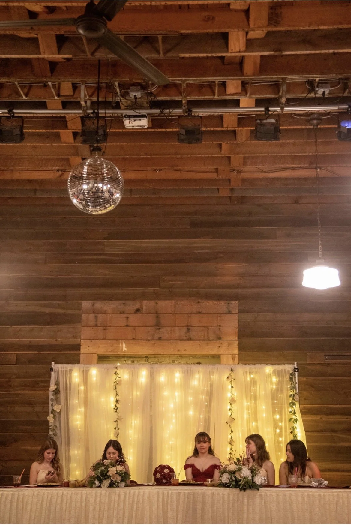 Four women sitting at a decorated table at a wedding reception, with a backdrop of yellow curtains and string lights, in a wood-paneled room with a chandelier and disco ball.