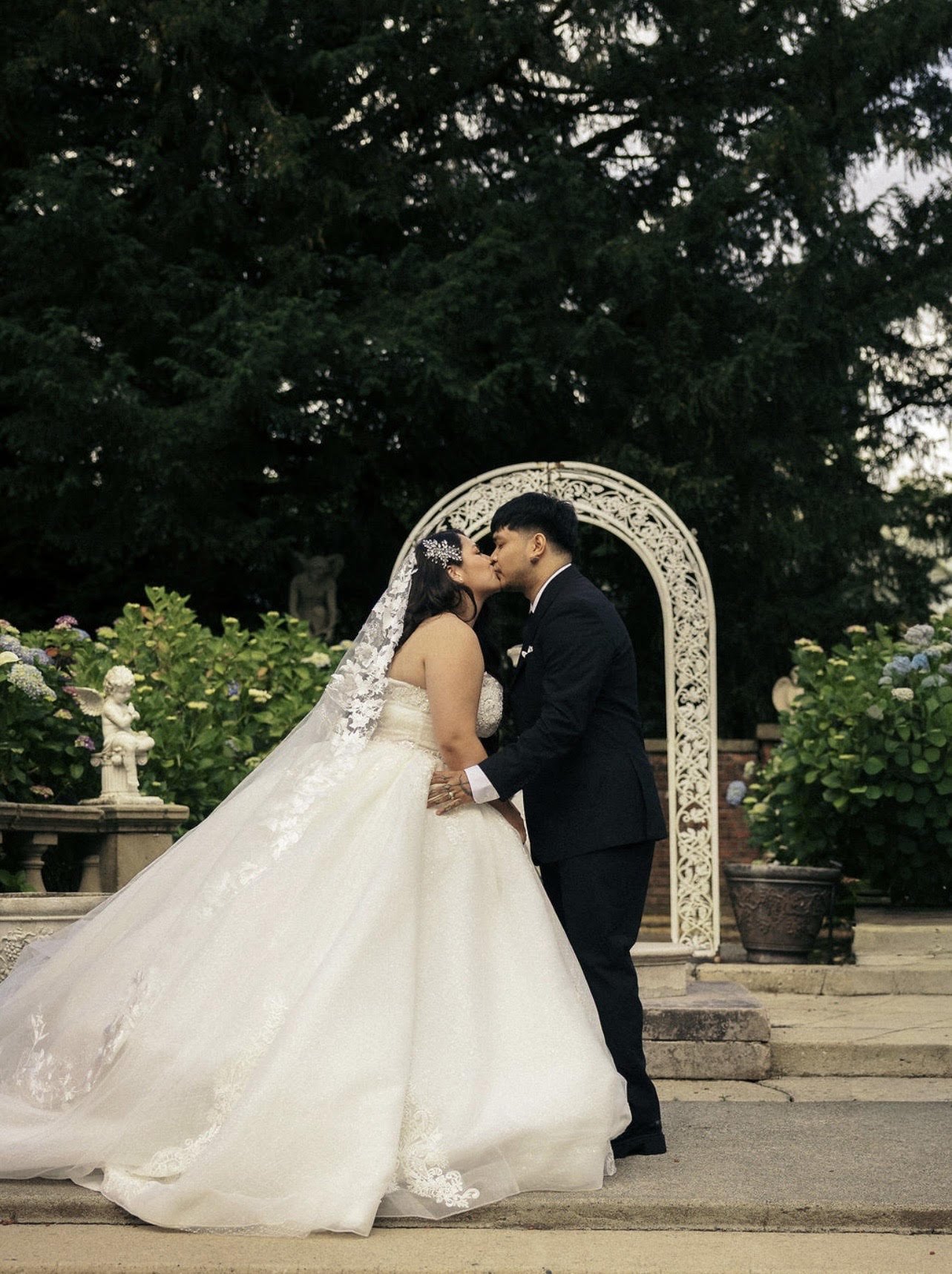 A bride and groom kiss during their wedding ceremony outdoors, surrounded by potted plants and flowers with a decorative arch in the background.