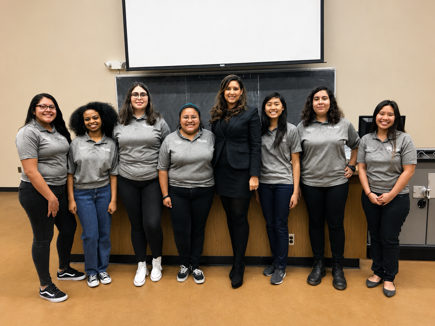 Group photo of nine women standing in front of a blackboard and a large white screen in a classroom. Eight women are wearing gray polo shirts and one woman in the center is wearing a black blazer and skirt. The women are smiling.