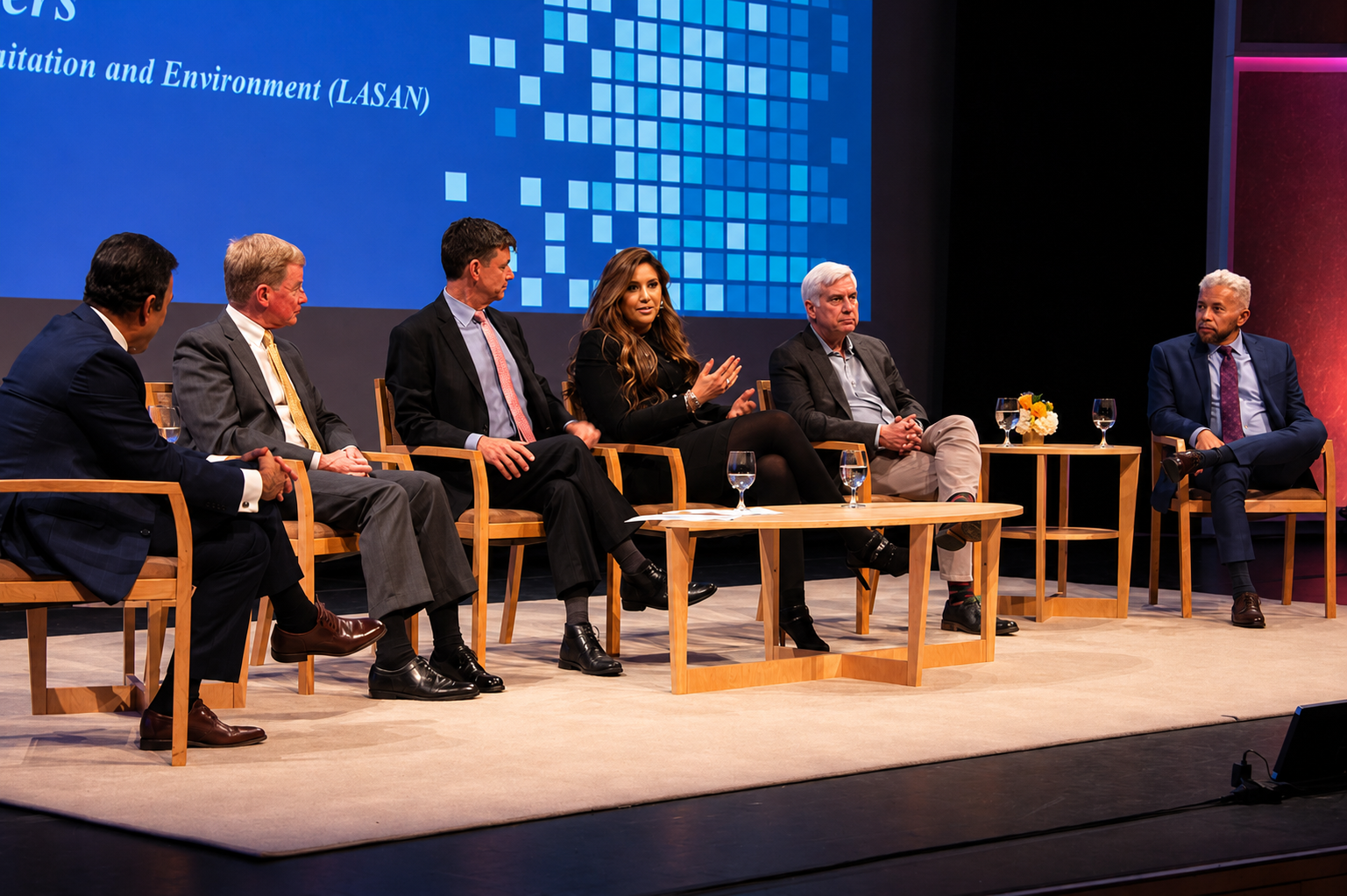 A panel of six people seated on stage during a discussion, with a large screen behind them displaying text.