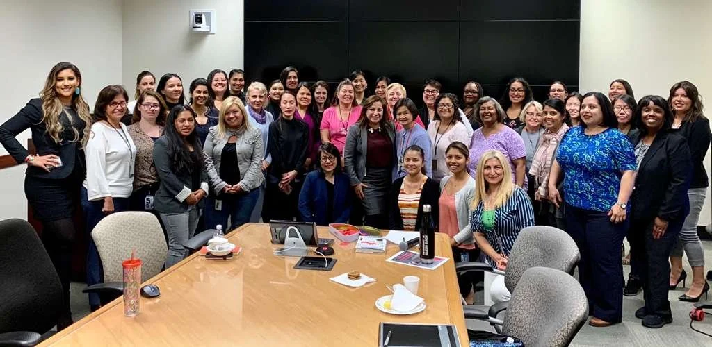 Group of women gathered in a conference room, posing for a photo around a large table, some seated and some standing, with a black wall in the background.