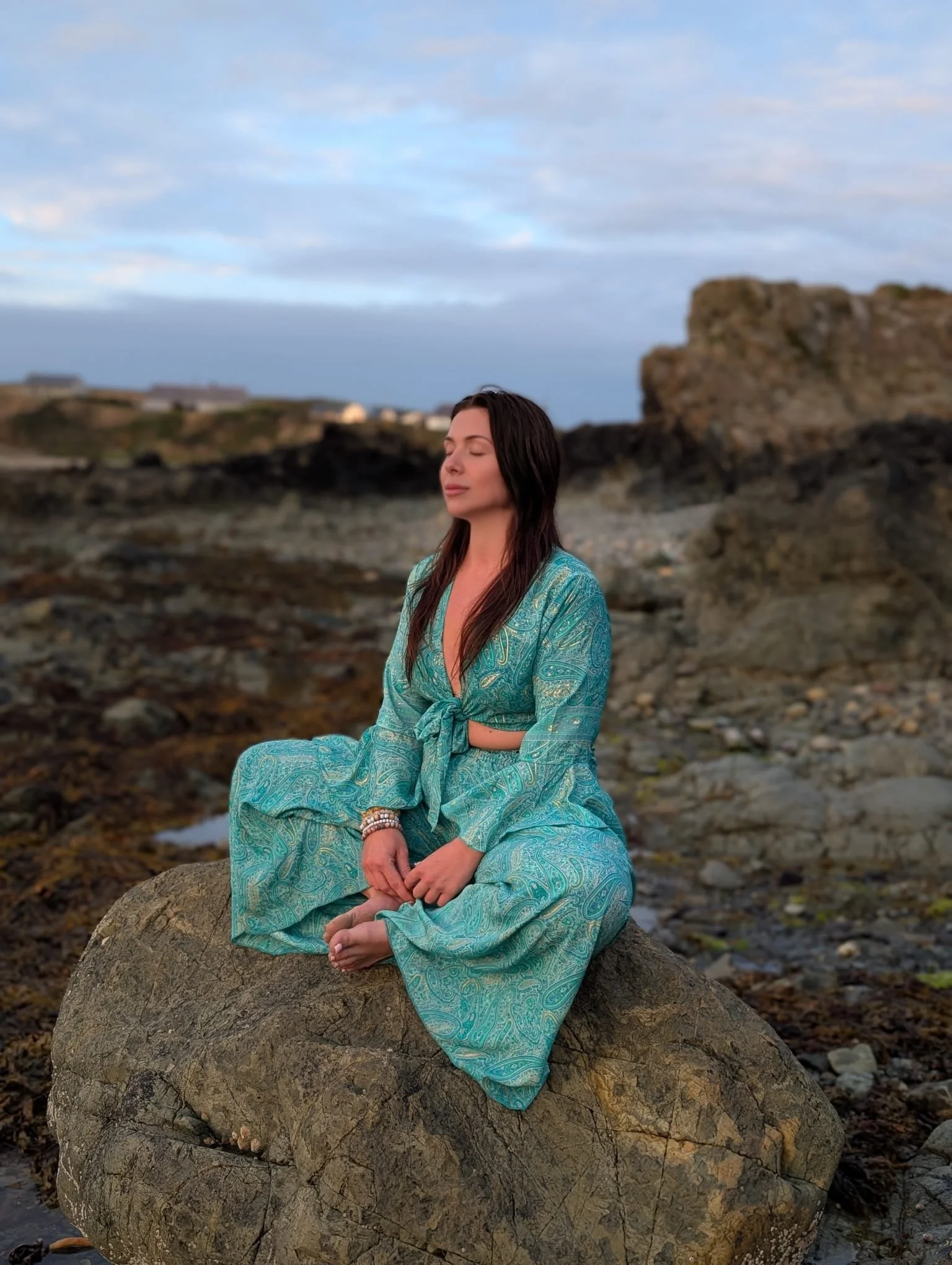 A woman with long dark hair in a teal patterned dress sitting on a large rock on a rocky beach with a cloudy sky in the background.