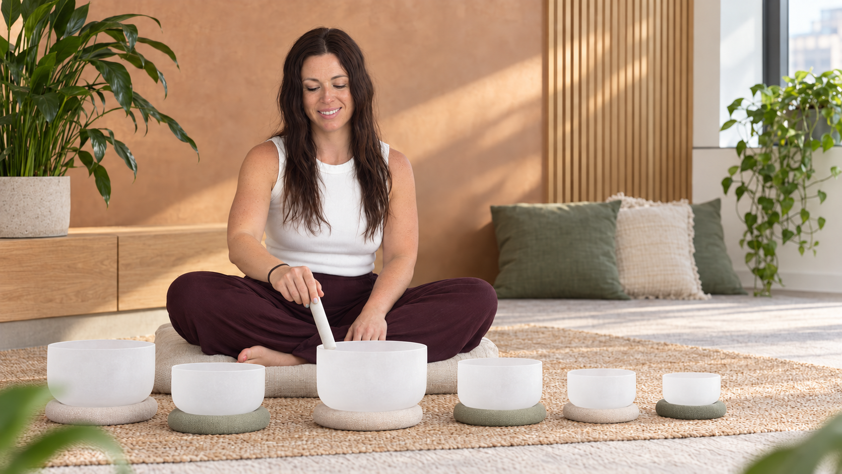 A woman sitting cross-legged on a rug, playing crystal singing bowls in a modern, cozy room with wooden accents and large windows, surrounded by houseplants.