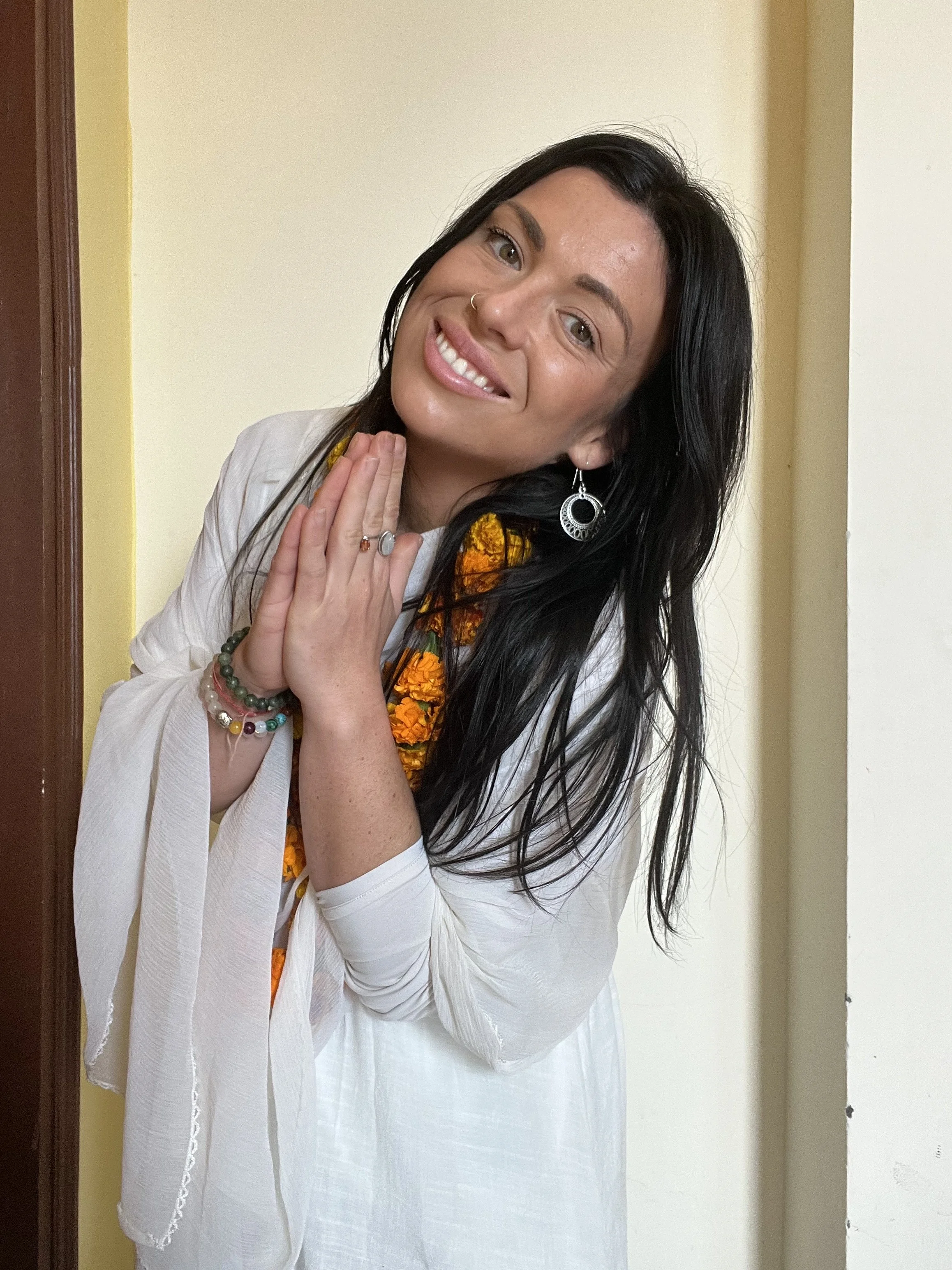 A woman with long black hair, wearing a white outfit, and jewelry including earrings, bracelets, and rings, smiling and holding her hands together near her face, standing against a plain wall.