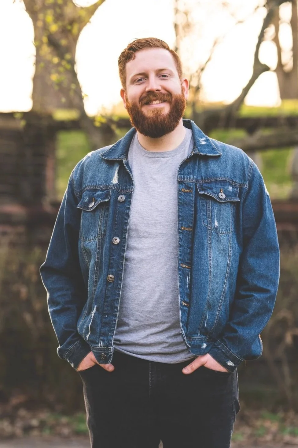 A man with red hair and a beard smiling outdoors, wearing a denim jacket and a gray T-shirt, standing in front of trees.
