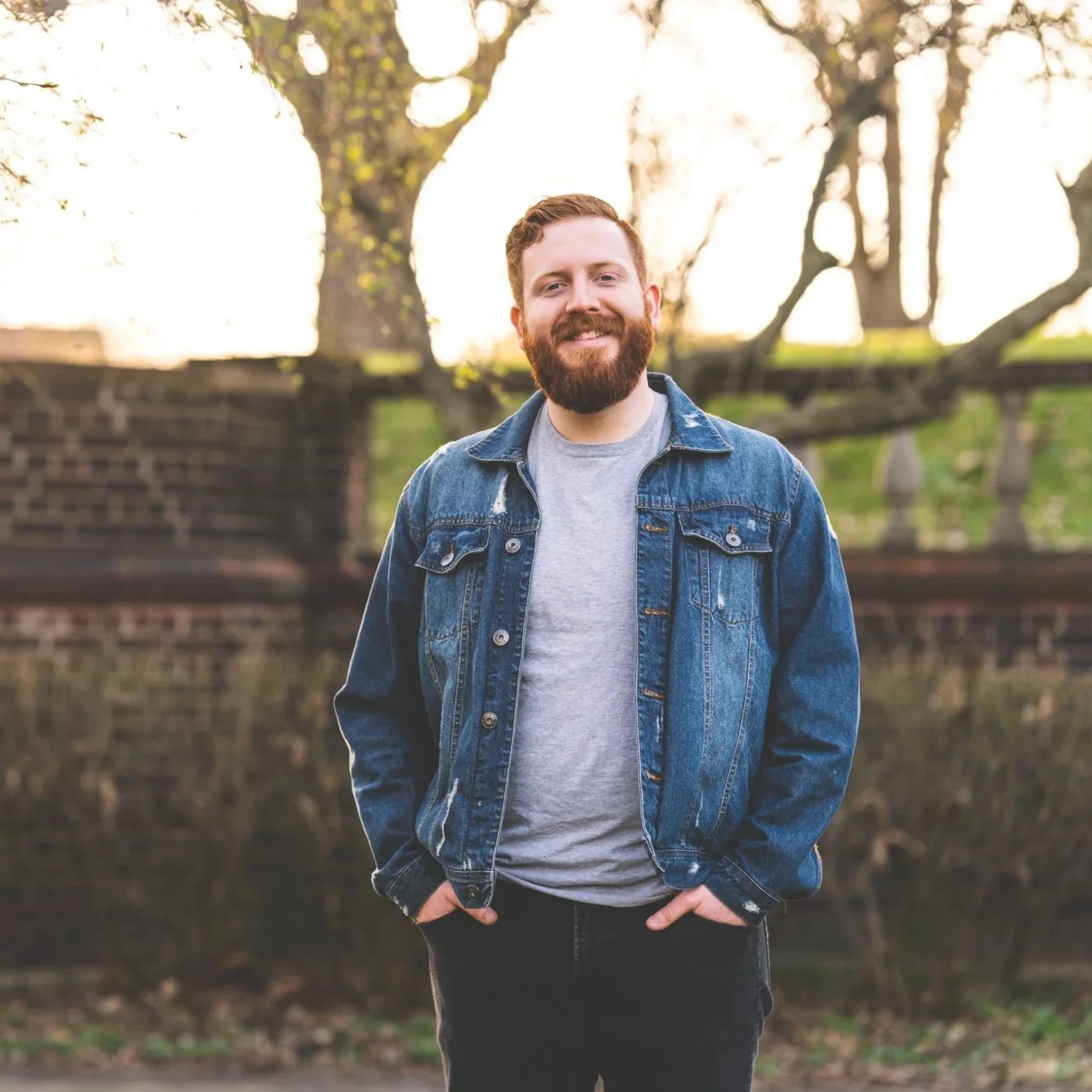 A man with a beard and short hair smiling while standing outdoors in front of a tree with branches and leaves. He's wearing a denim jacket over a gray t-shirt, with hands in his pockets, during what appears to be sunset or sunrise.