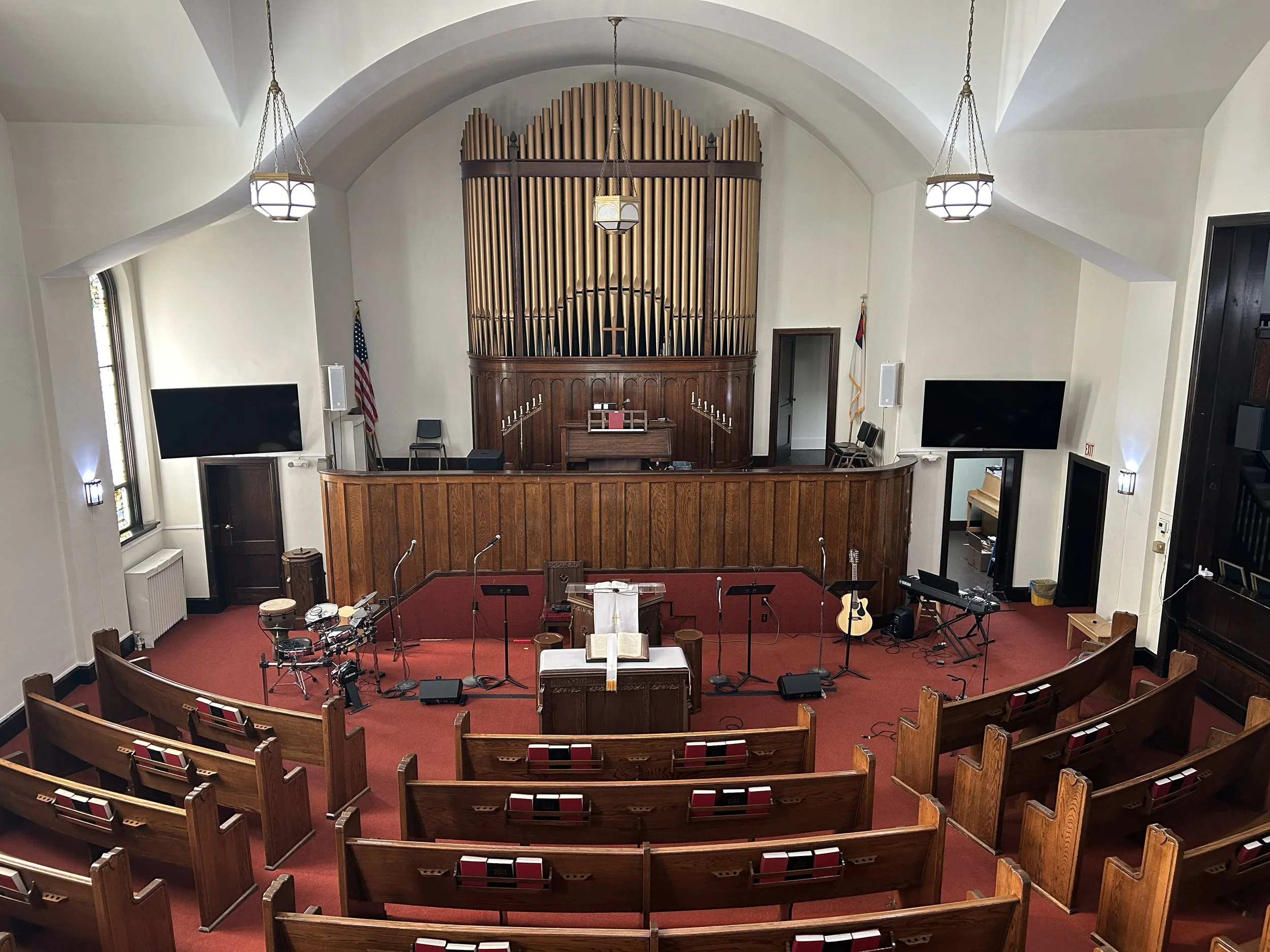 Inside view of a church sanctuary with wooden choir loft, musical instruments, microphones, large black screens, wooden pews with red cushions, and a pipe organ at the front. Flags are positioned on the left and right sides.