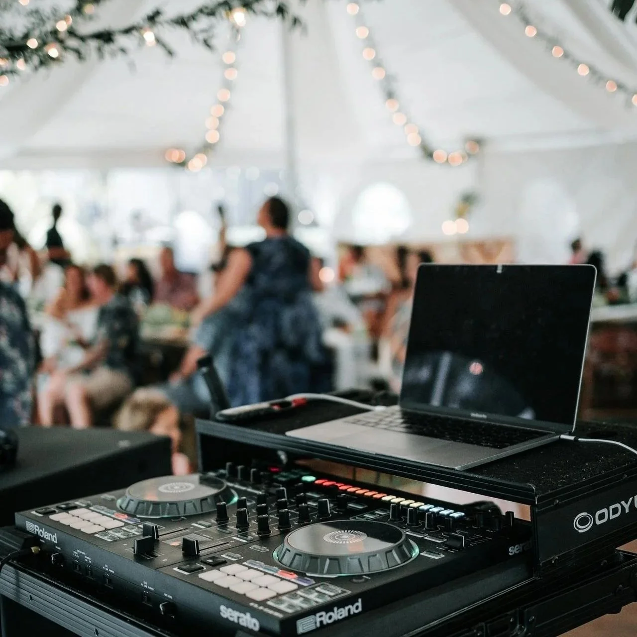 DJ equipment with a laptop and mixer at an event with people in the background under string lights.