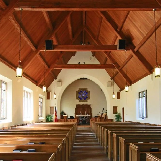 Interior of a church with wooden pews, high vaulted wooden ceiling, and a stage at the front with musical instruments and a flag.
