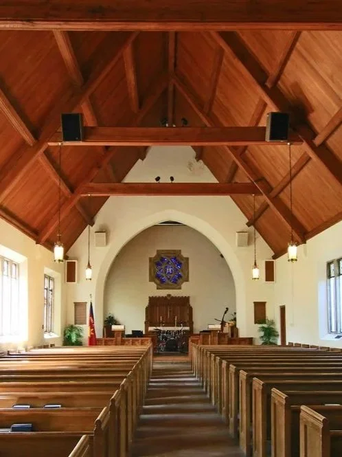 View of the interior of a church with wooden pews, a high wooden ceiling, and an altar at the front with a decorative wall hanging.