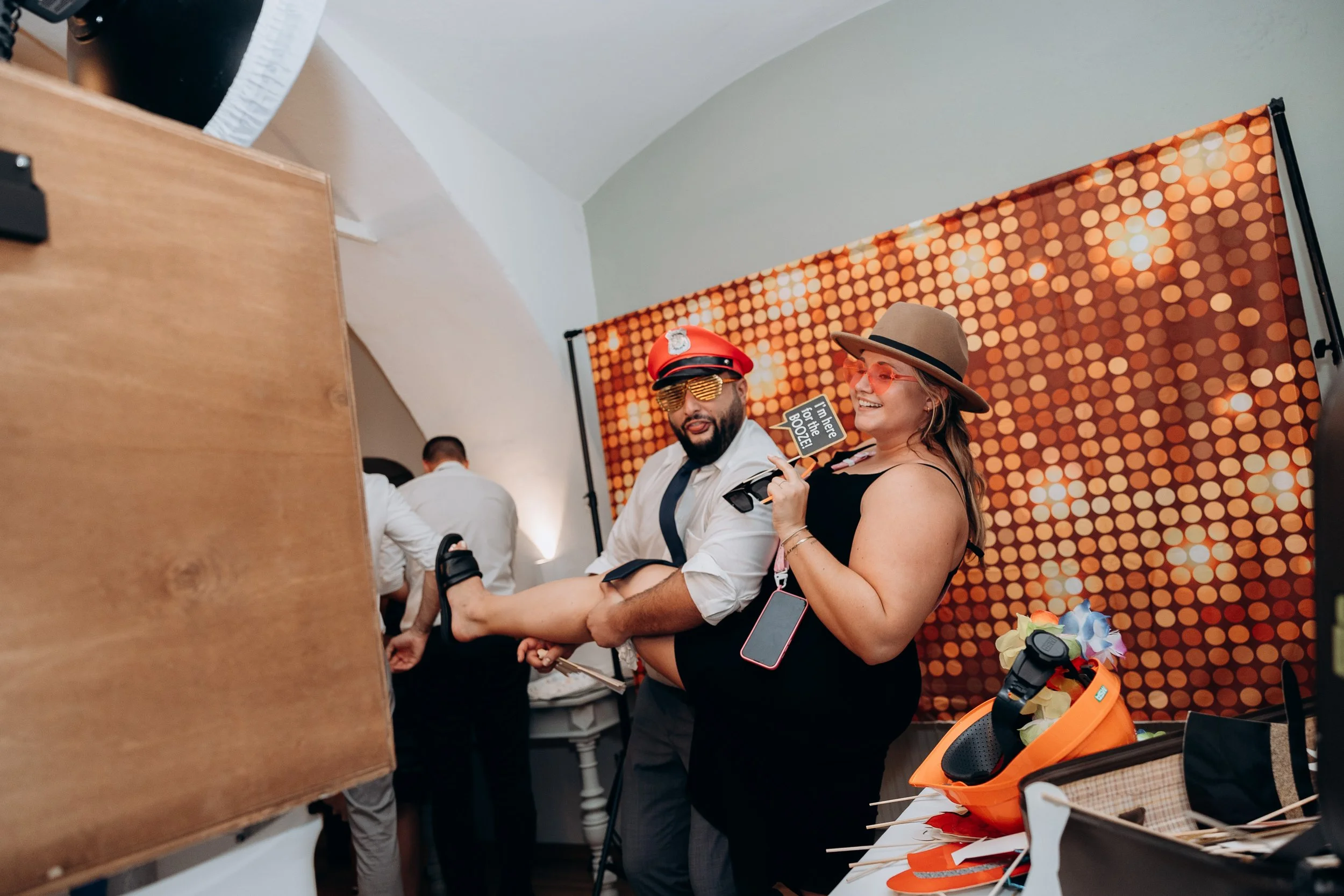 Zwei Personen tragen Fotohüten und Sonnenbrillen mit lila Rahmen und posieren vor einem orangefarbenen, doppelseitigen Lichtpunkt-Hintergrund. Sie lachen und stehen in einem Raum mit weißen Wänden, auf dem Tisch liegen Blumen und Fotorequisiten.
