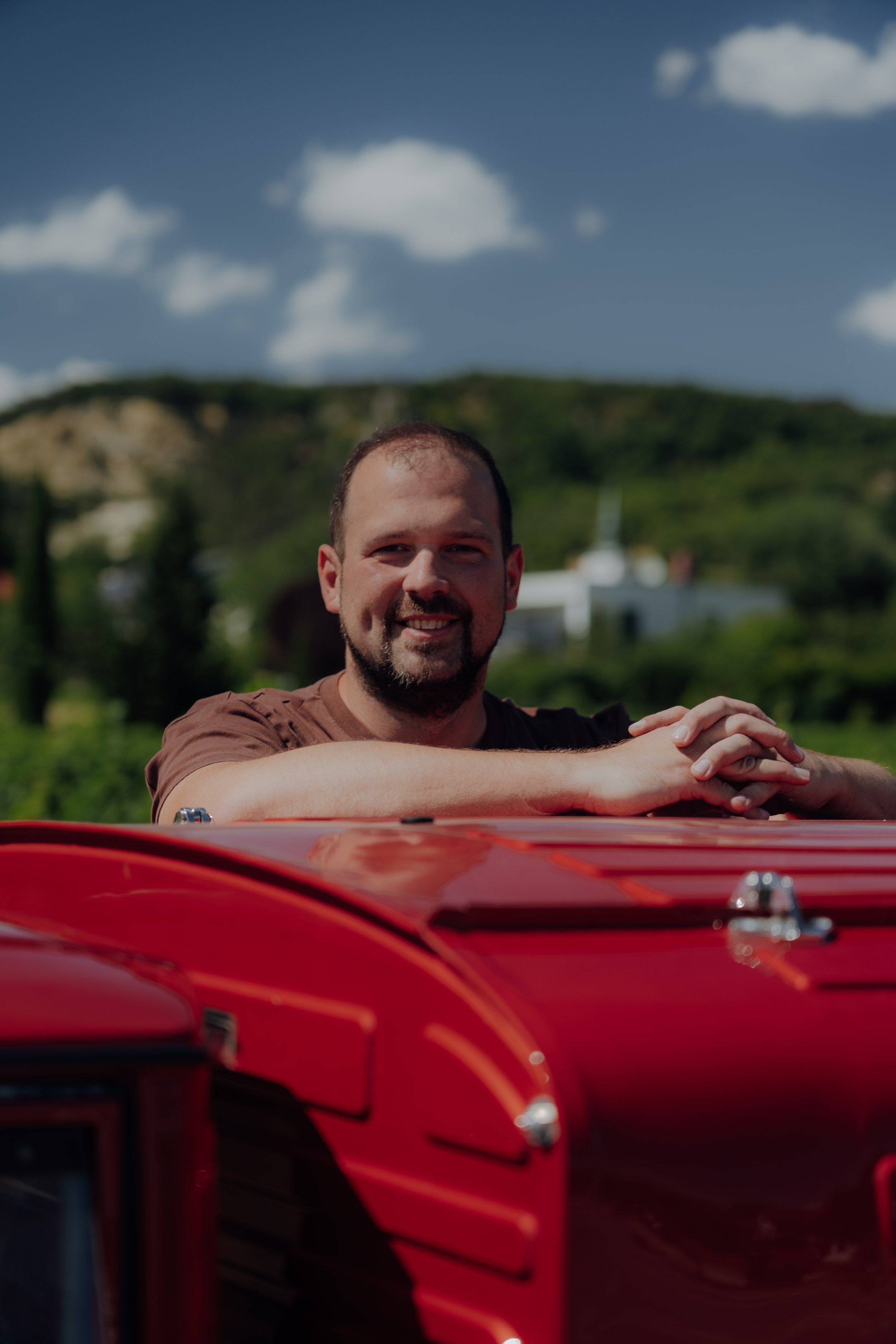 Ein lachender Mann lehnt auf dem Auto, das im Vordergrund sichtbar ist, während er in einer ländlichen Umgebung mit Hügeln und einem blauen Himmel mit einigen Wolken steht.