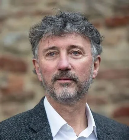 A middle-aged man with curly gray hair and a beard, wearing a white shirt and gray blazer, standing outdoors in front of a brick wall.