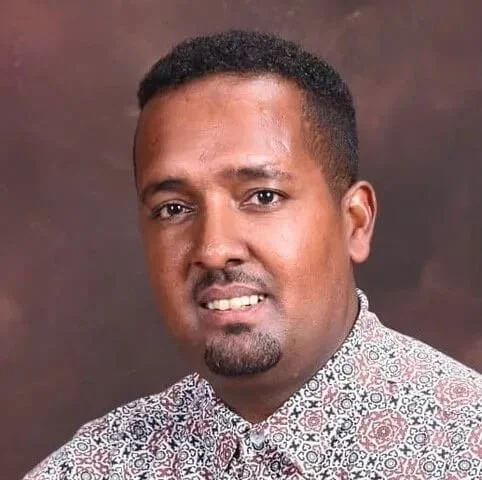 Headshot of a man with short, curly hair and a goatee, wearing a patterned shirt, smiling against a plain brown background.