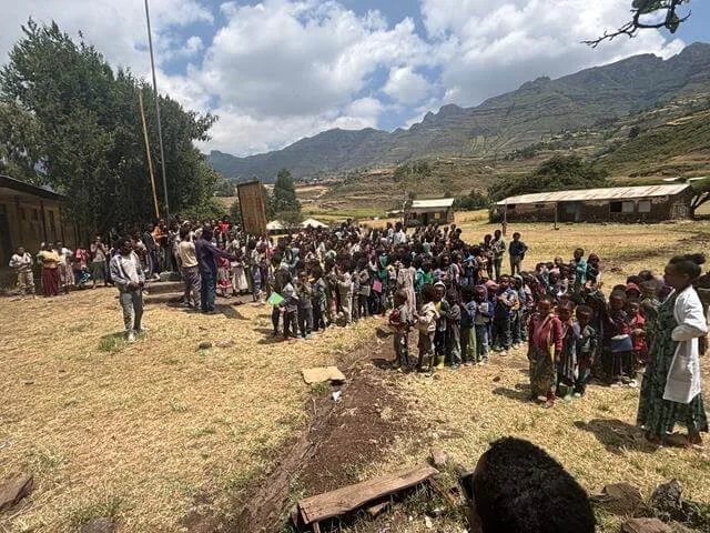 A large group of children and adults gathered outdoors in a rural area with mountains in the background. The scene appears to be a community gathering or event, with some people standing in lines and others mingling.