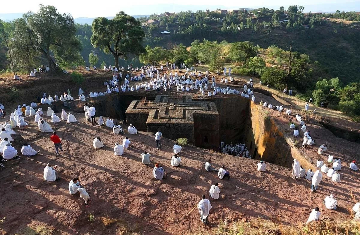 Numerous people dressed in white are gathered on a hillside around a large, dark structure resembling a temple or monument, with trees and a landscape in the background.
