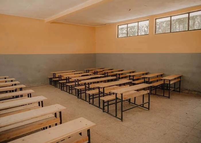 Empty classroom with wooden desks arranged in rows, a yellow wall, gray lower wall, a window with multiple panes, and a tiled floor.