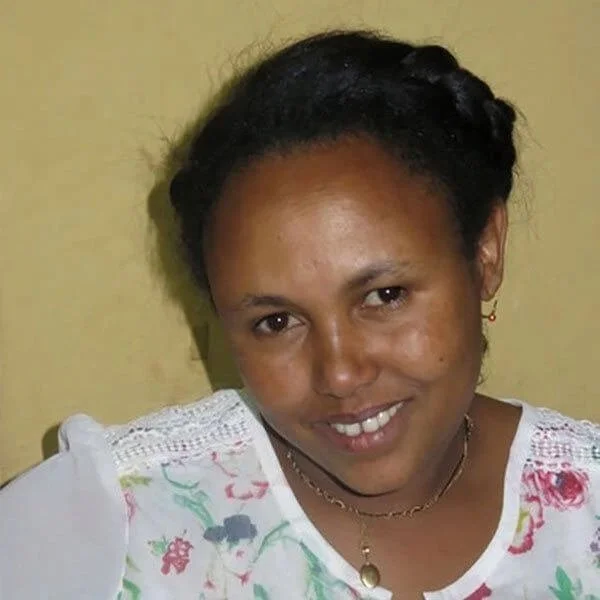 A woman with dark hair styled in braids, wearing a white top with colorful floral patterns, a gold necklace, and earrings, smiling at the camera against a yellow wall.