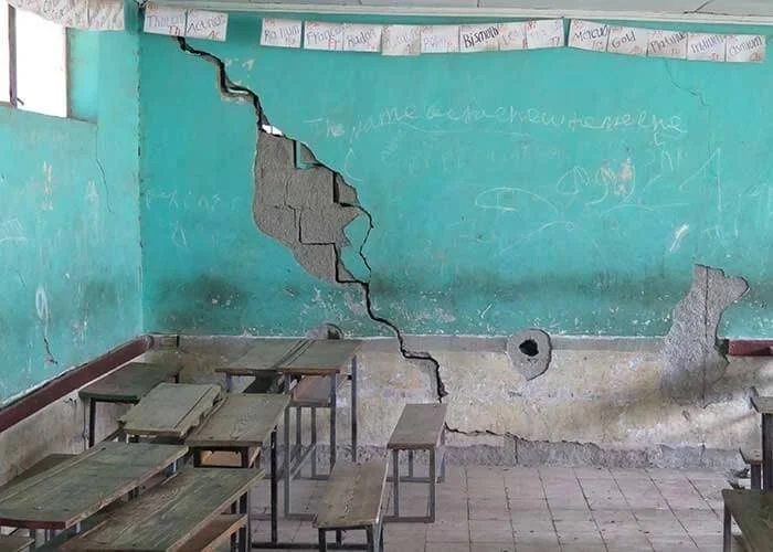 A classroom with a large crack in the turquoise wall, exposing concrete behind it. There are chalkboards, desks, and a banner hanging with written names or words.