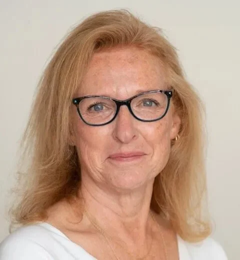 A woman with shoulder-length blonde hair wearing black glasses and a white top, smiling slightly against a plain light background.