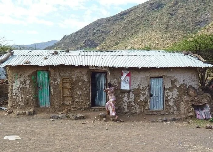 A woman in a skirt standing outside a mud house with three doors, one green, one blue, and one gray, against a backdrop of hills and a partly cloudy sky.