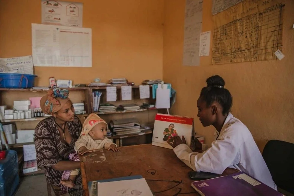 A woman holding a child sitting at a desk with a healthcare professional in a white coat in a modest office setting.