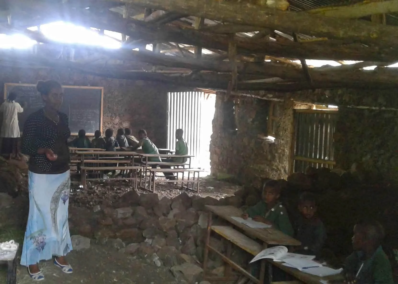 A classroom inside a rustic, partially open building with students sitting at benches and a teacher at the front. The structure has stone walls and a wooden roof with daylight coming through gaps.