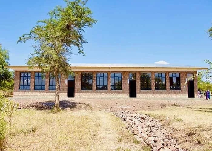 A single-story brick building with multiple large windows and a flat roof, surrounded by a dry grassy area and trees, under a clear blue sky.