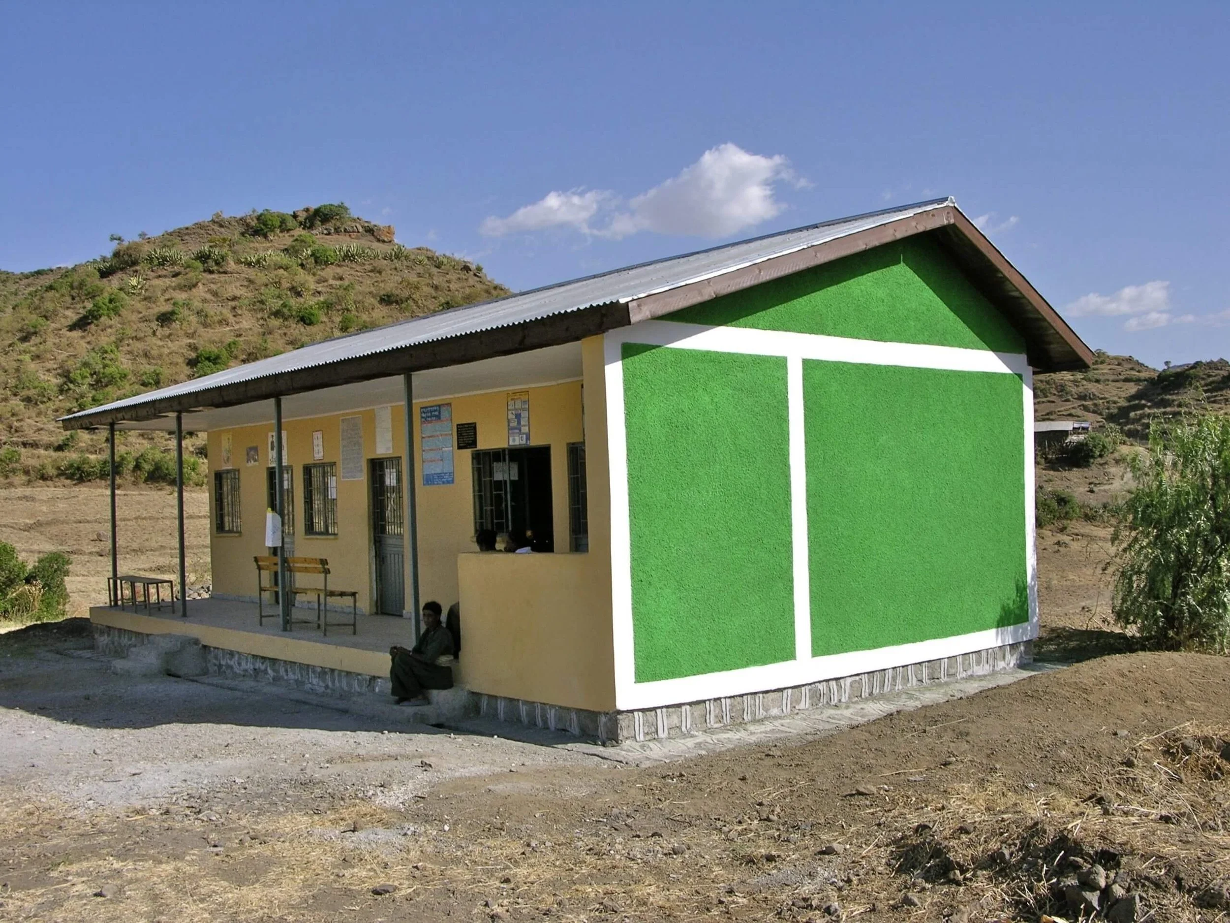 A small building with green and yellow walls, a metal roof, and a porch supported by black poles. Two people are sitting outside, and the building is set against a background of rolling hills and a bright blue sky with some clouds.